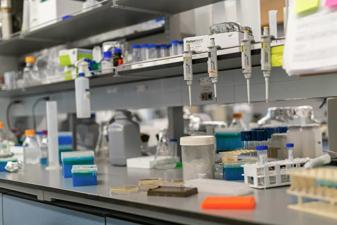 Zatłoczony blat laboratorium biomedycznego z pipetami, butelkami z odczynnikami, probówkami, statywami i pojemnikami, oddający atmosferę skupionej pracy naukowej.